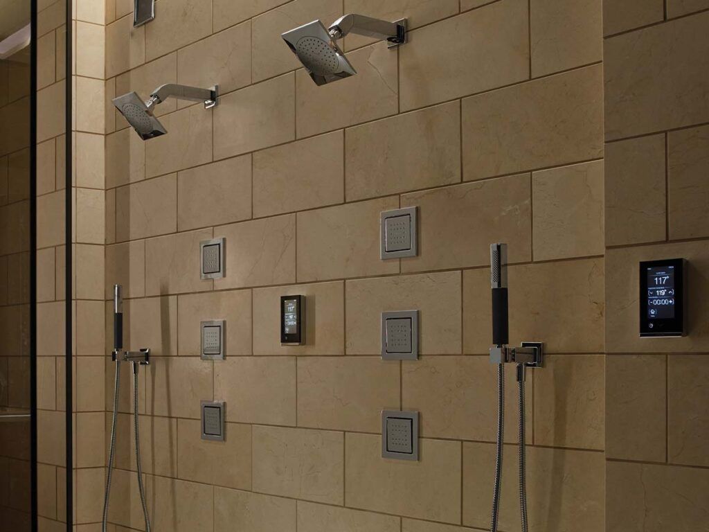 A moody shower with beige tile and square motifs, featuring body sprays, handshowers, rainshowers, and a digital valve sysyem.