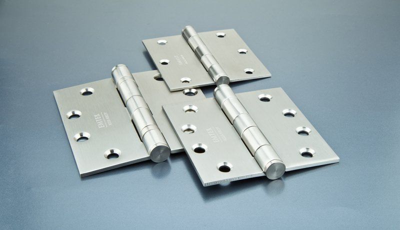 Three stainless steel Emtek hinges on a gray background.