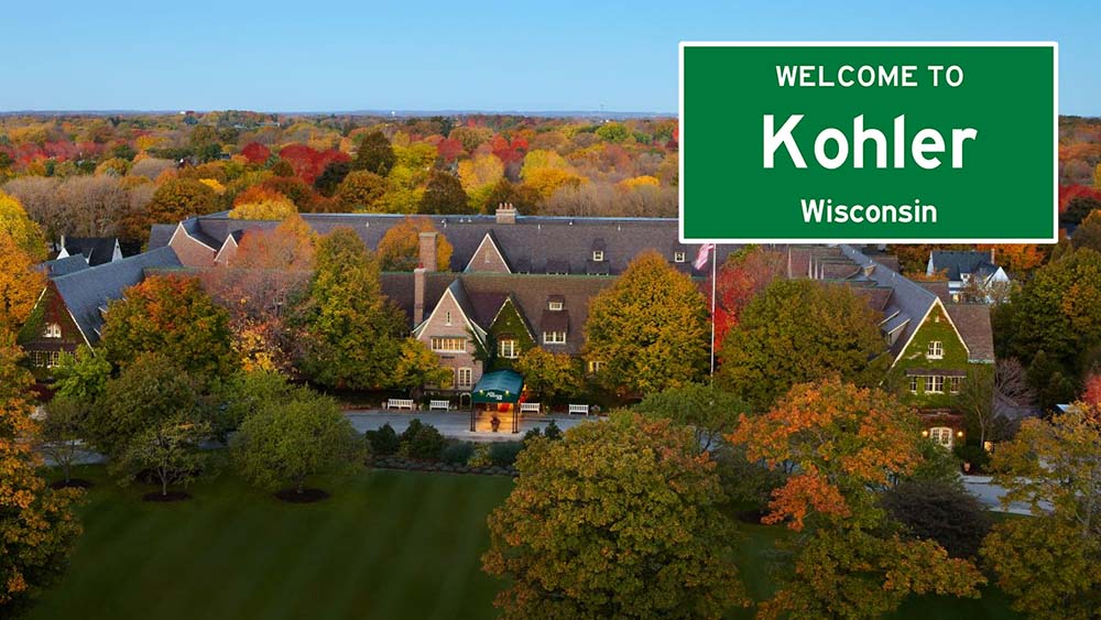 an aerial view of the kohler american inn surounded by fall foliage, a green highway style sign says welcome to kohler wisconsin