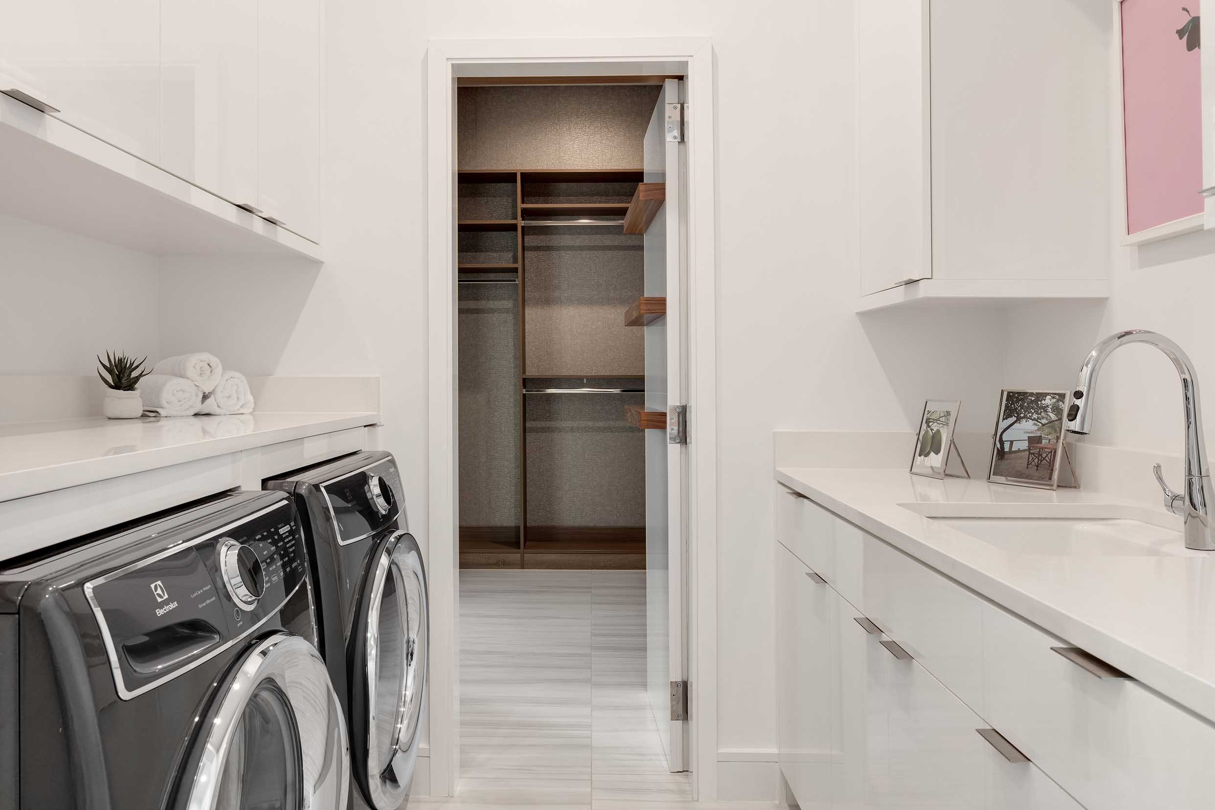A compact, clean, white galley-style laundry room that opens into a large clothing closet with custom cabinetry.
