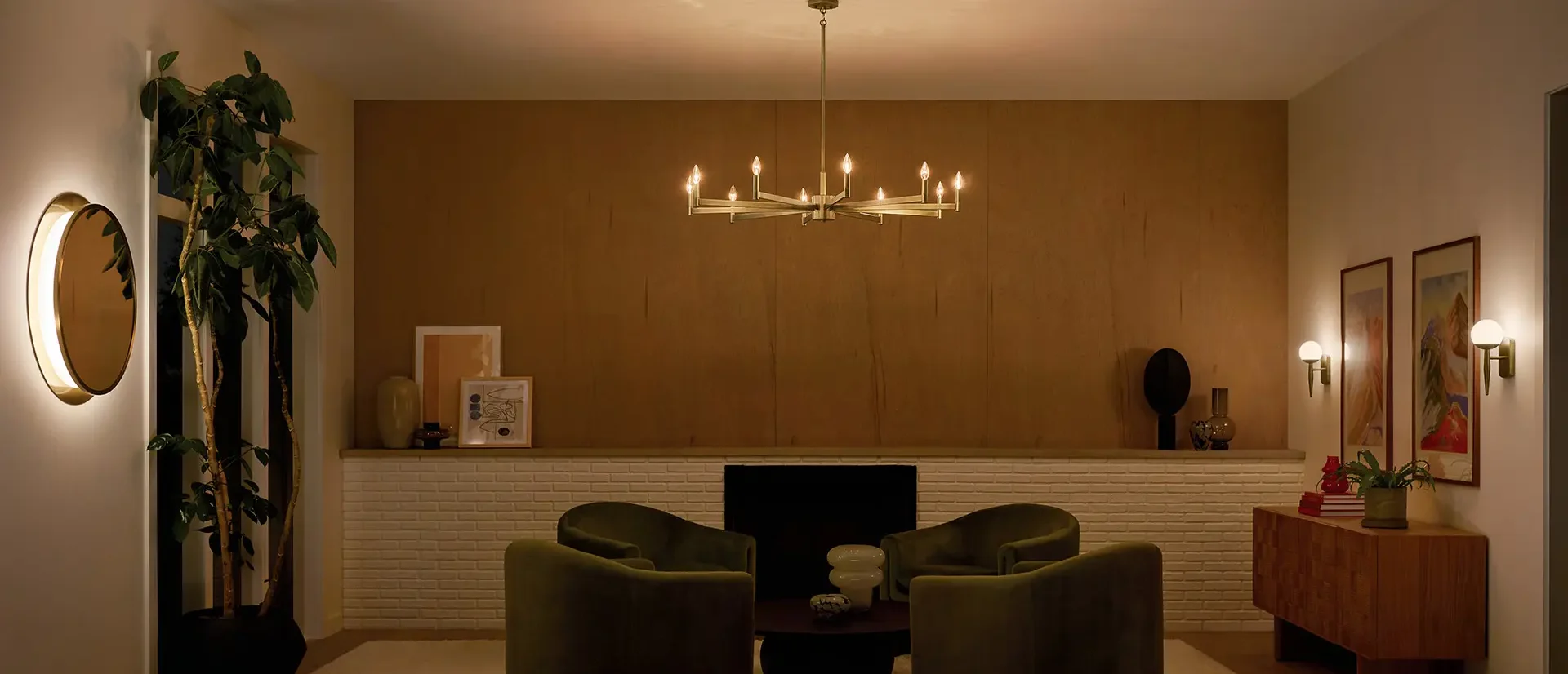Cozy living room at night featuring a modern chandelier, green upholstered seating, and a white brick fireplace with a wooden mantle.