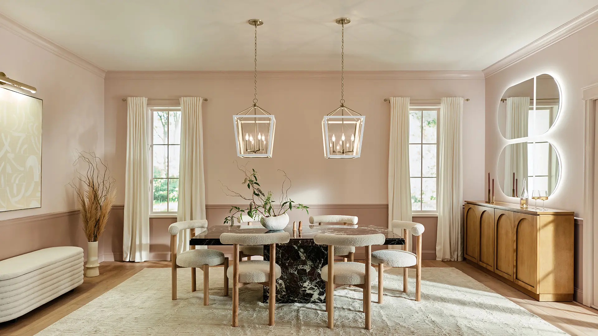 Elegant dining room in soft pink tones with a black marble table, cream upholstered chairs, and two geometric pendant lights. A curved-back mirror sits above a wooden sideboard.